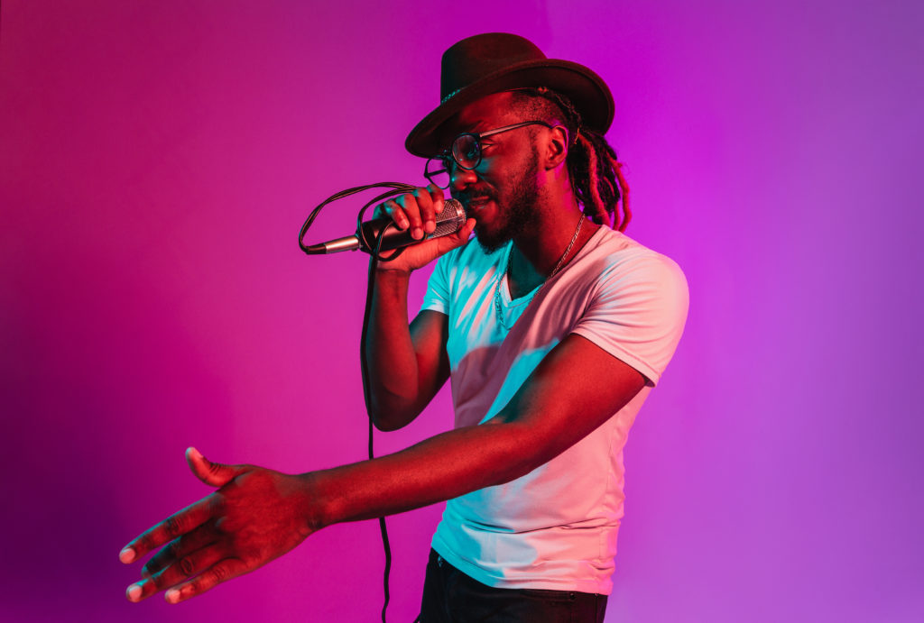 Young african-american jazz musician with microphone singing a song on purple studio background in trendy neon light. Concept of music, hobby, inspirness. Colorful portrait of joyful attractive artist.