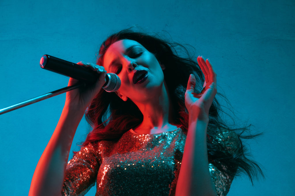 Caucasian female singer portrait isolated on blue studio background in neon light. Beautiful female model in bright dress with microphone. Concept of human emotions, facial expression, ad, music, art.